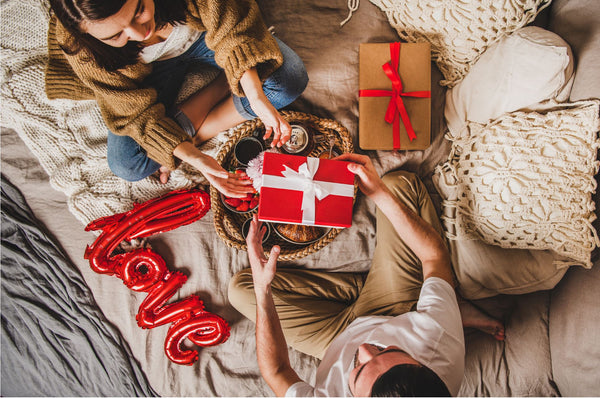 a male and female sitting on the bed with wrapped presents and a balloon which spells love